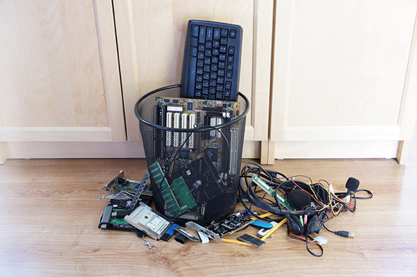 Poubelle de bureau remplie de matériel informatique usagé, avec d’autres équipements sur le parquet