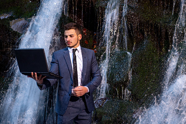 Jeune commercial en costume se tenant sous des chutes d’eau avec un PC portable durci étanche